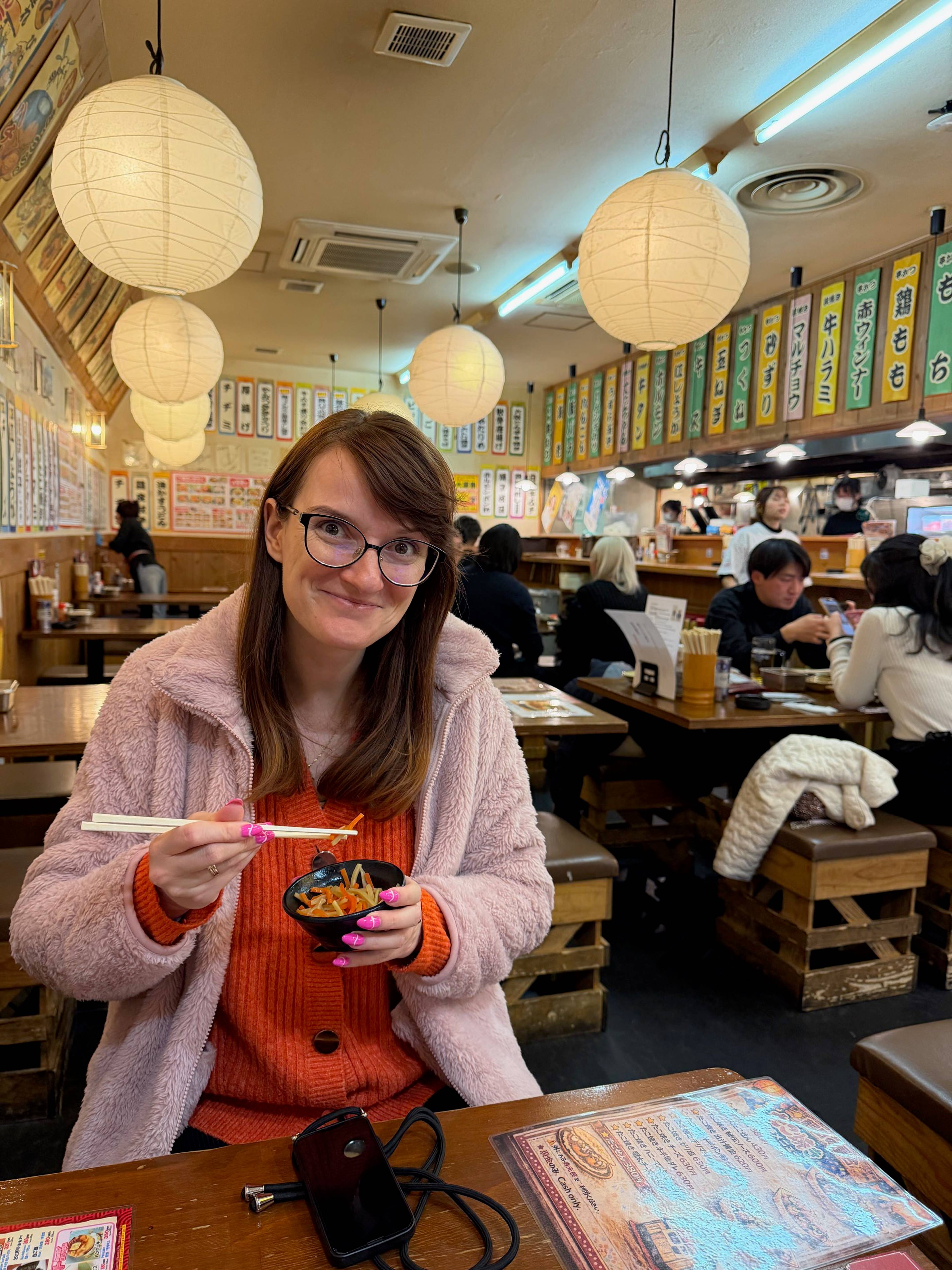 Kobieta siedzi przy stoliku w japońskiej restauracji i je posiłek pałeczkami, a wokół znajdują się wiszące papierowe lampiony, menu na ścianach i inni goście.