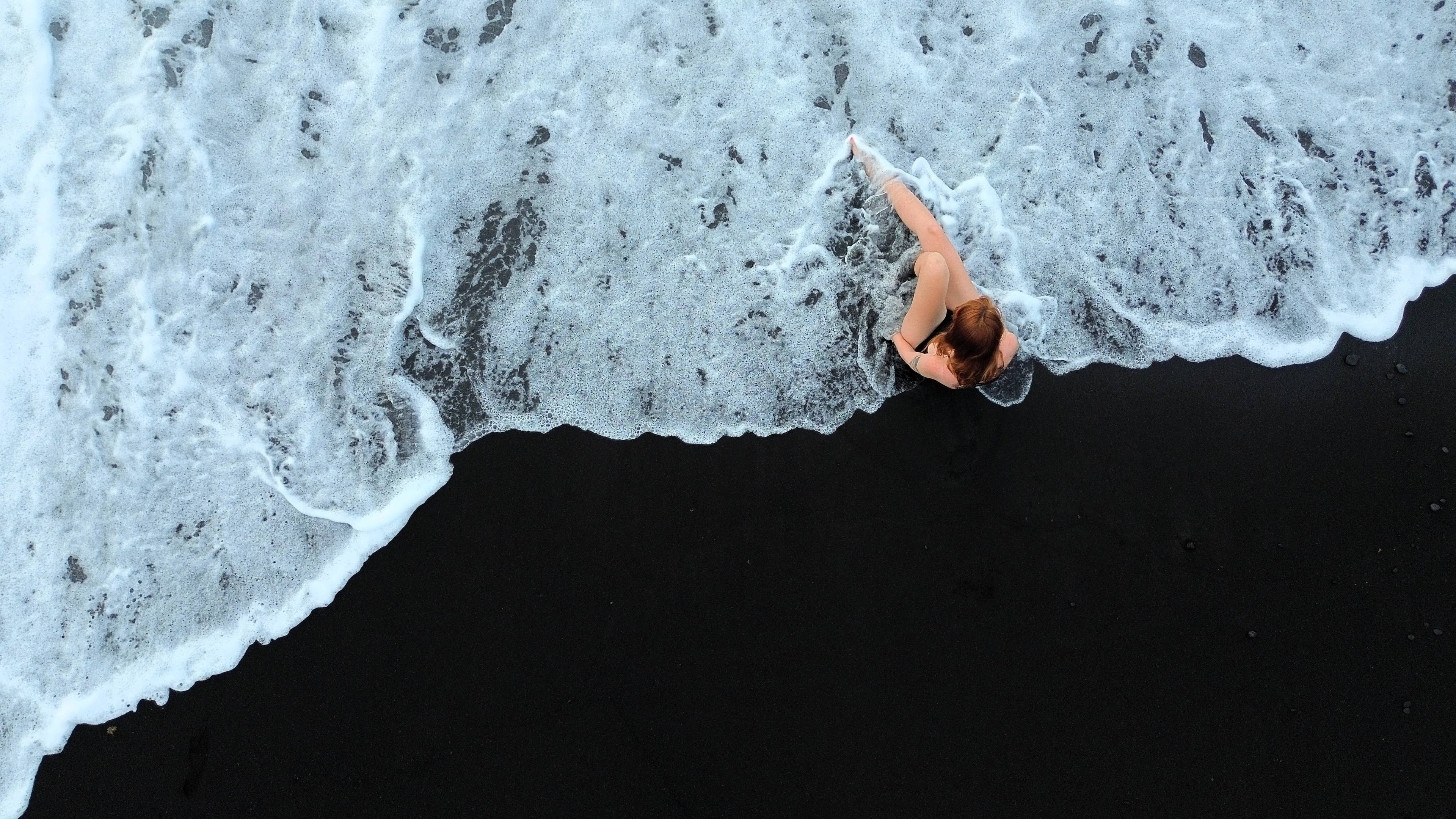 Rudowłosa dziewczyna siedzi na czarnym piasku na plaży i obmywa ją biała piana z morskiej fali