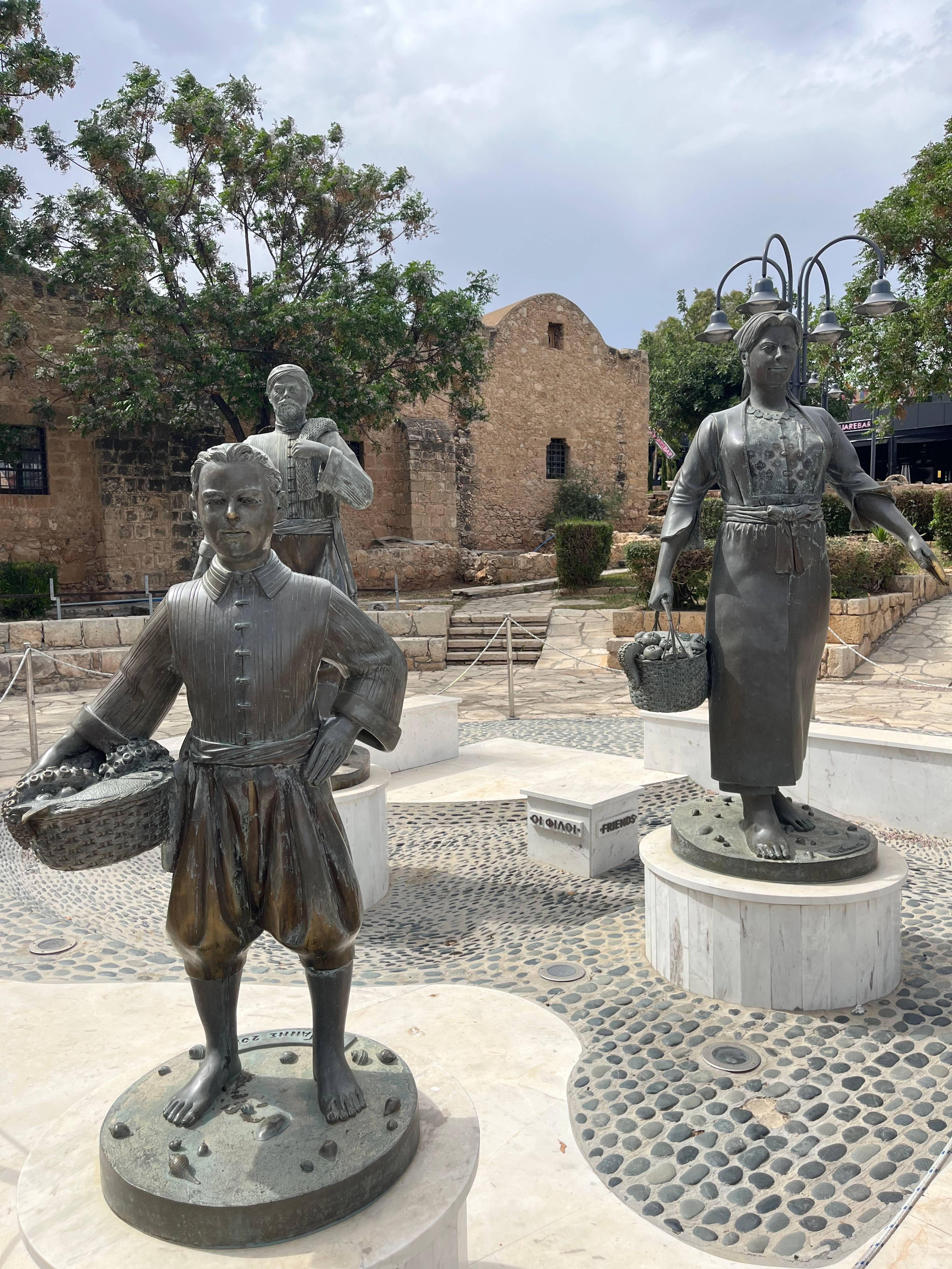 Metalowe rzeźby przedstawiające tradycyjnych mieszkańców z koszami, ustawione na kamiennej posadzce w centrum Ayia Napa na Cyprze.