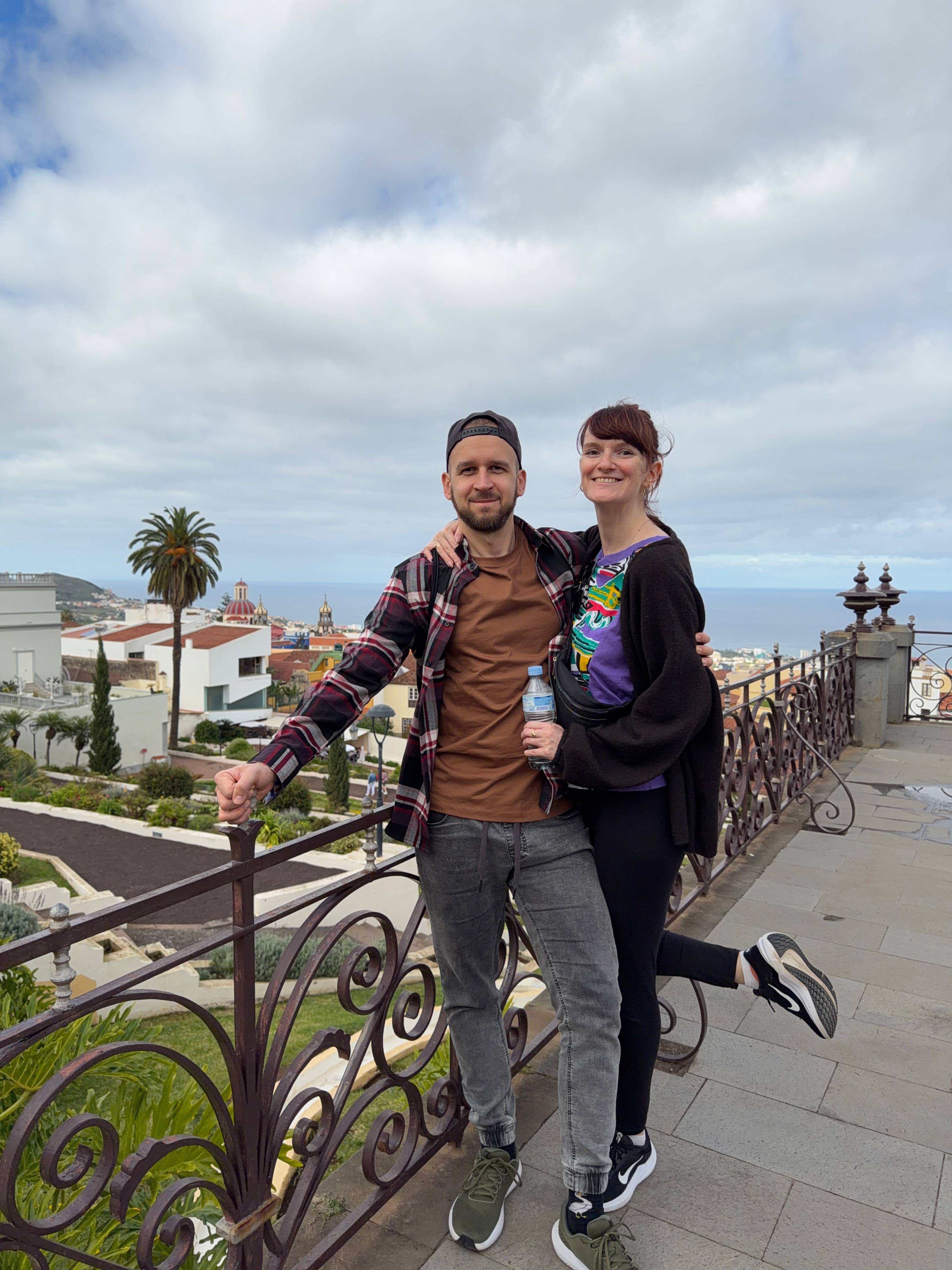 Dwie osoby pozują na tarasie z kutą balustradą z malowniczym widokiem na La Orotava, palmę i ocean w tle.