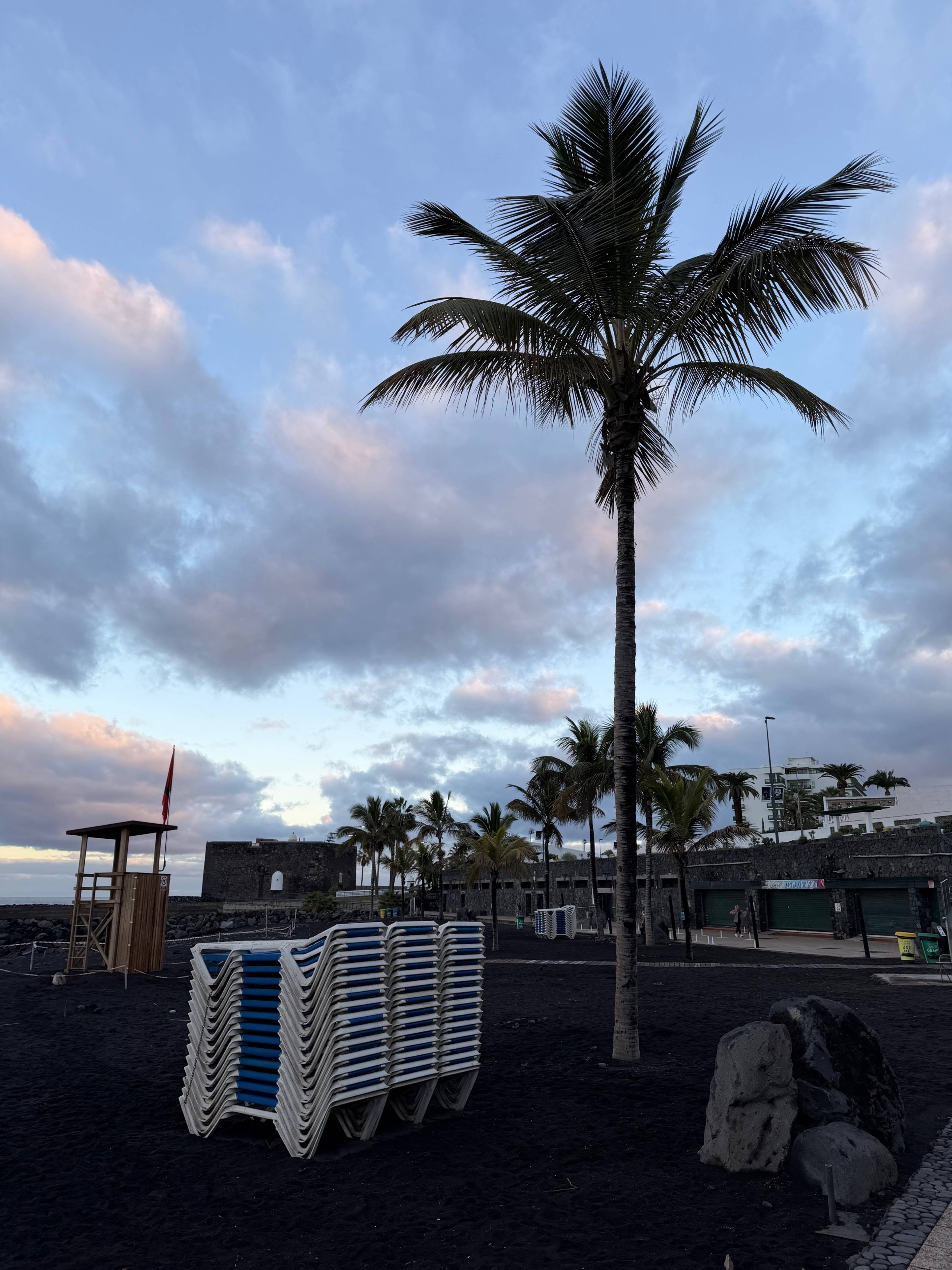 Plaża z czarnym piaskiem, złożone leżaki jeden na drugim, drewniana butka ratownika i palma.