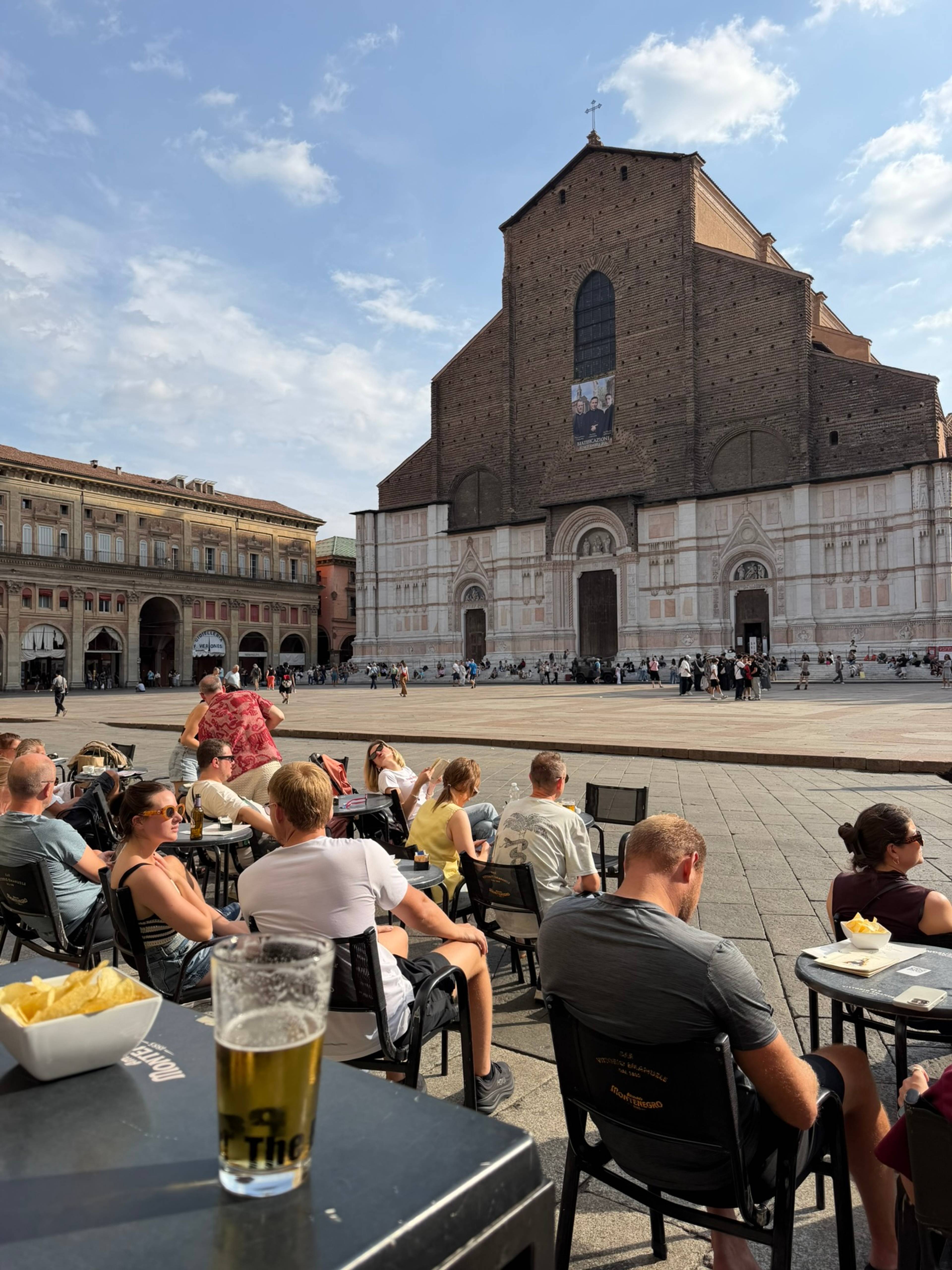 Monumentalna fasada Bazyliki San Petronio z ceglaną górną częścią i jasną marmurową dolną częścią góruje nad placem pełnym ludzi.