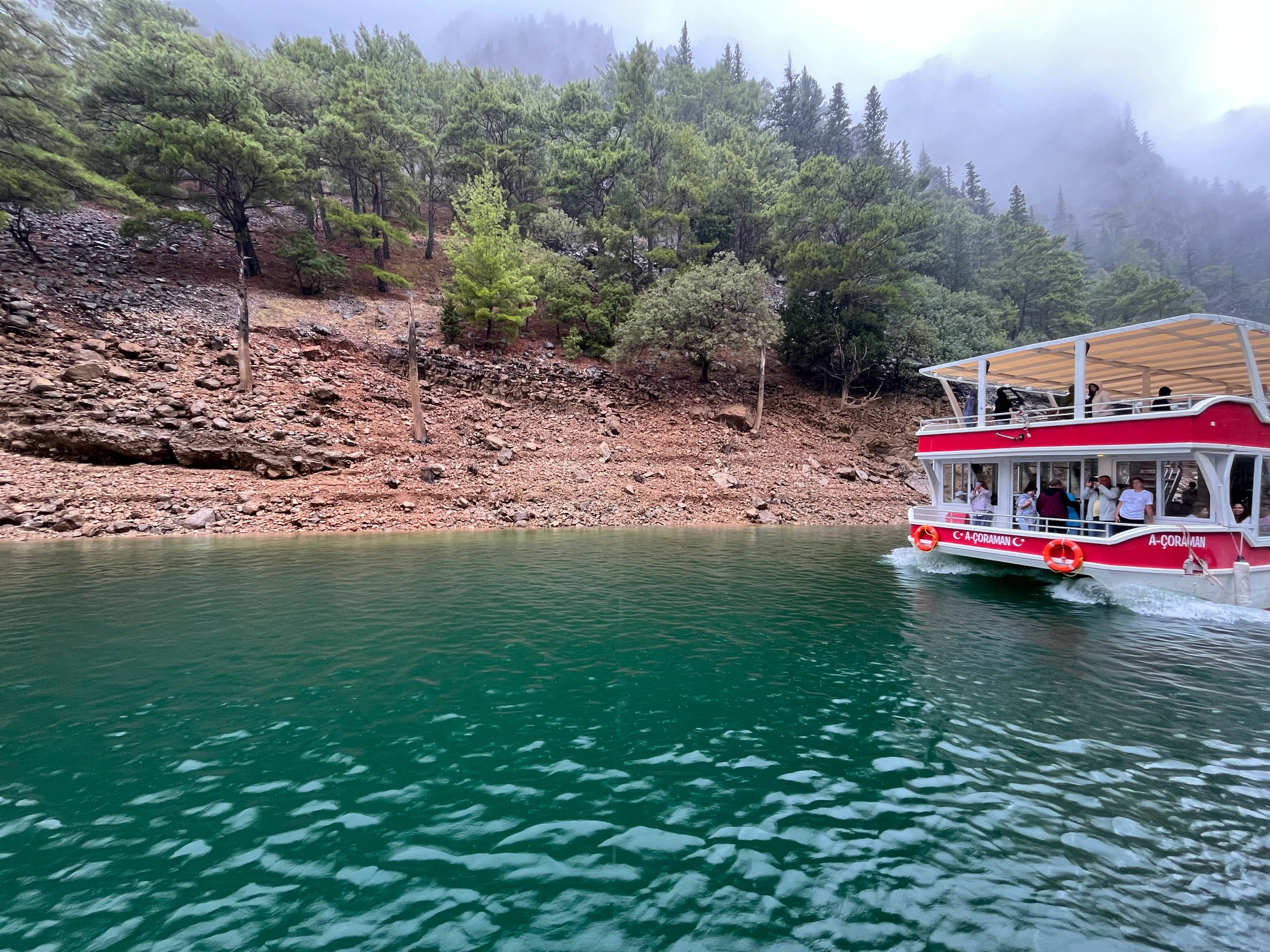 Statek turystyczny na zielonkawej wodzie w pobliżu skalistego brzegu porośniętego gęstymi sosnami, w tle mgła nad górami.