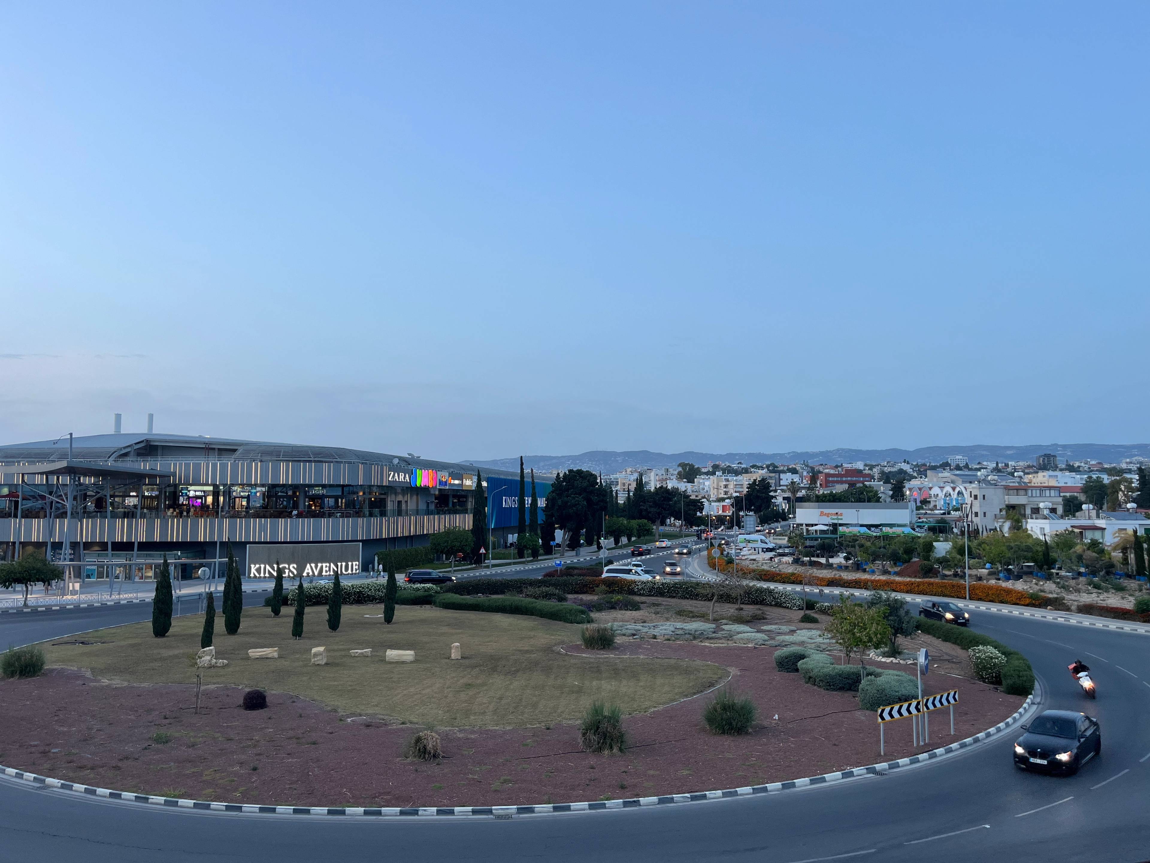 Rondo z zagospodarowanym środkiem, otoczone drogami z pojazdami; w tle nowoczesny budynek z napisem 'KING'S AVENUE' i sklepami, a dalej drzewa i inne zabudowania pod czystym niebem.
