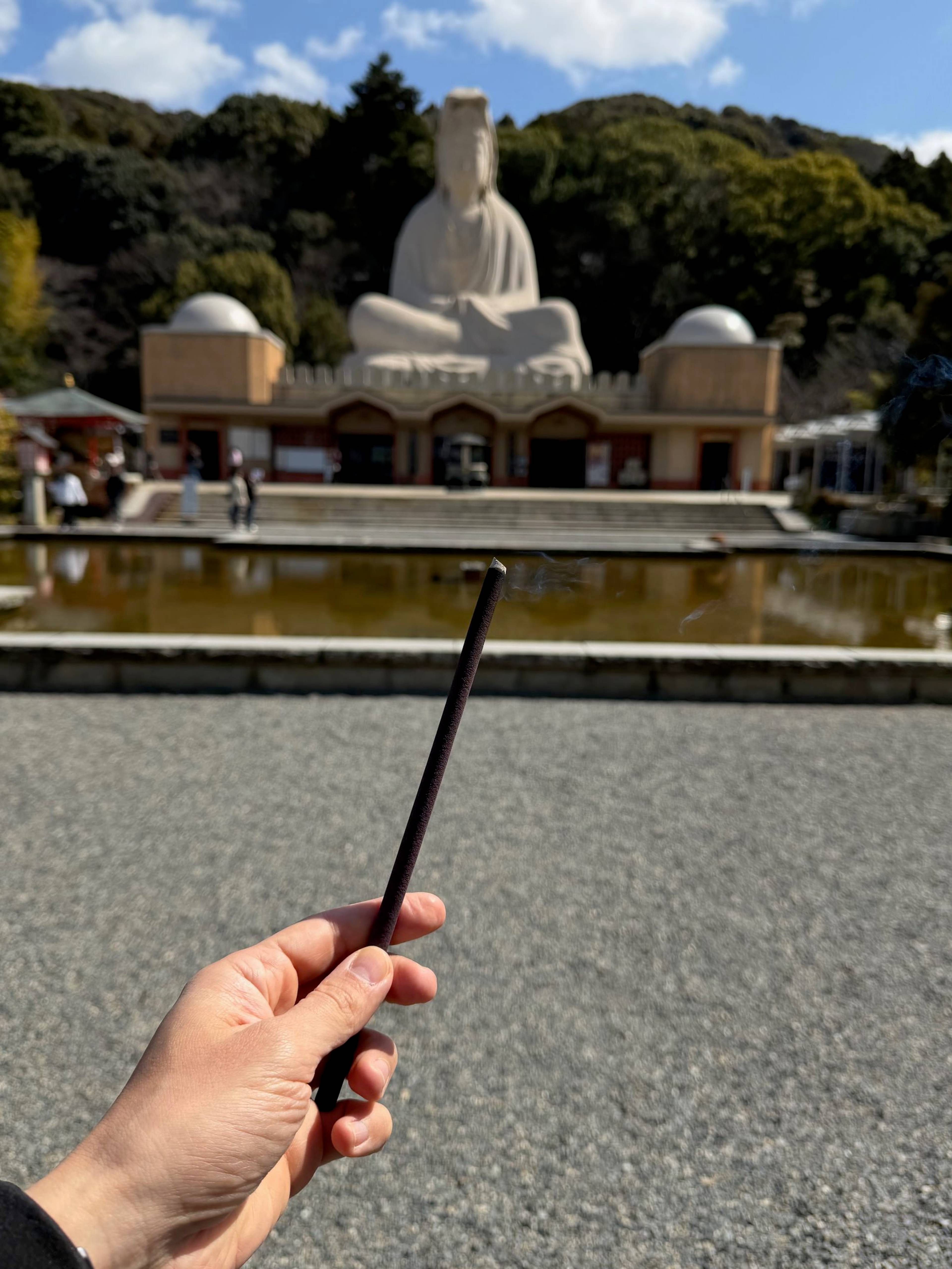 Dłoń trzyma tlące się kadzidełko, a w tle znajduje się dziedziniec świątyni z dużym posągiem i zbiornikiem z wodą.