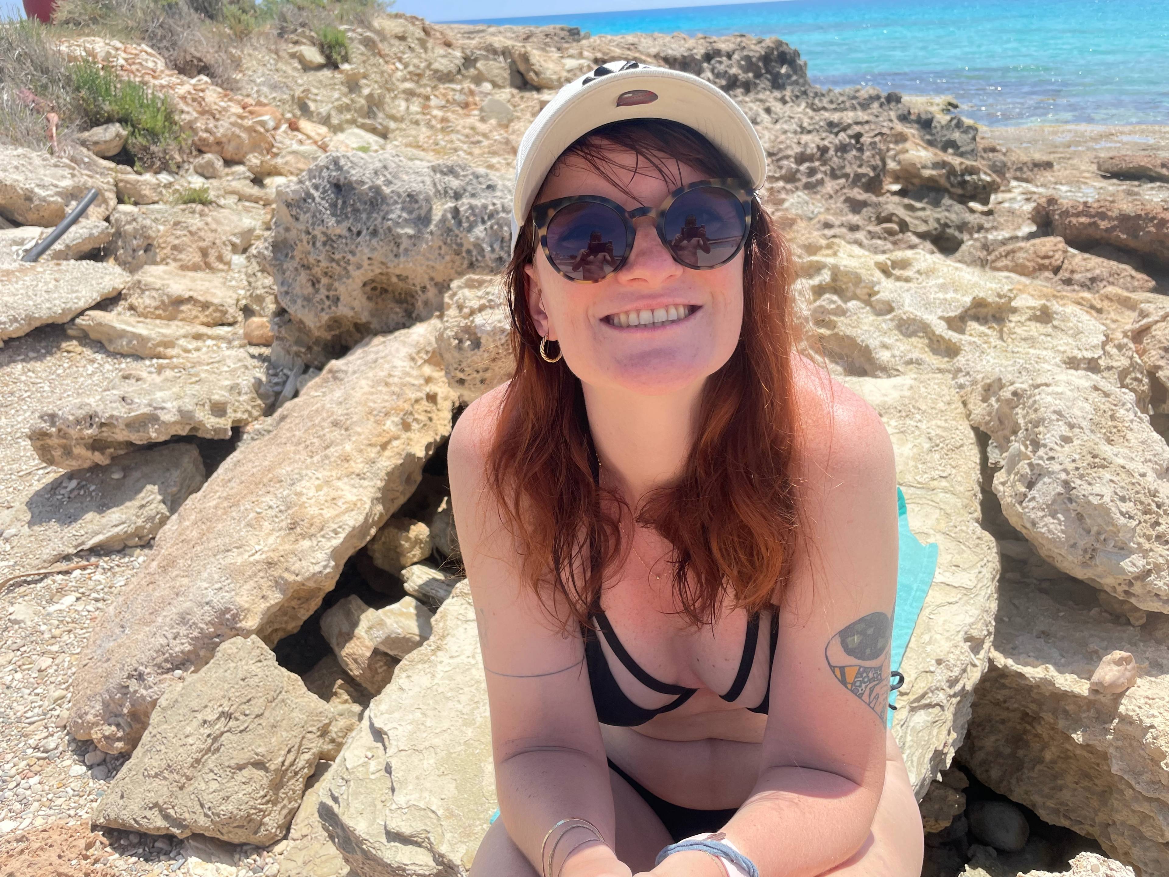 Uśmiechnięta, ruda dziewczyna w kostiumie kąpielowym, czapce z daszkiem i okularach przeciwsłonecznych, siedzi na skałach na plaży na Cyprze.