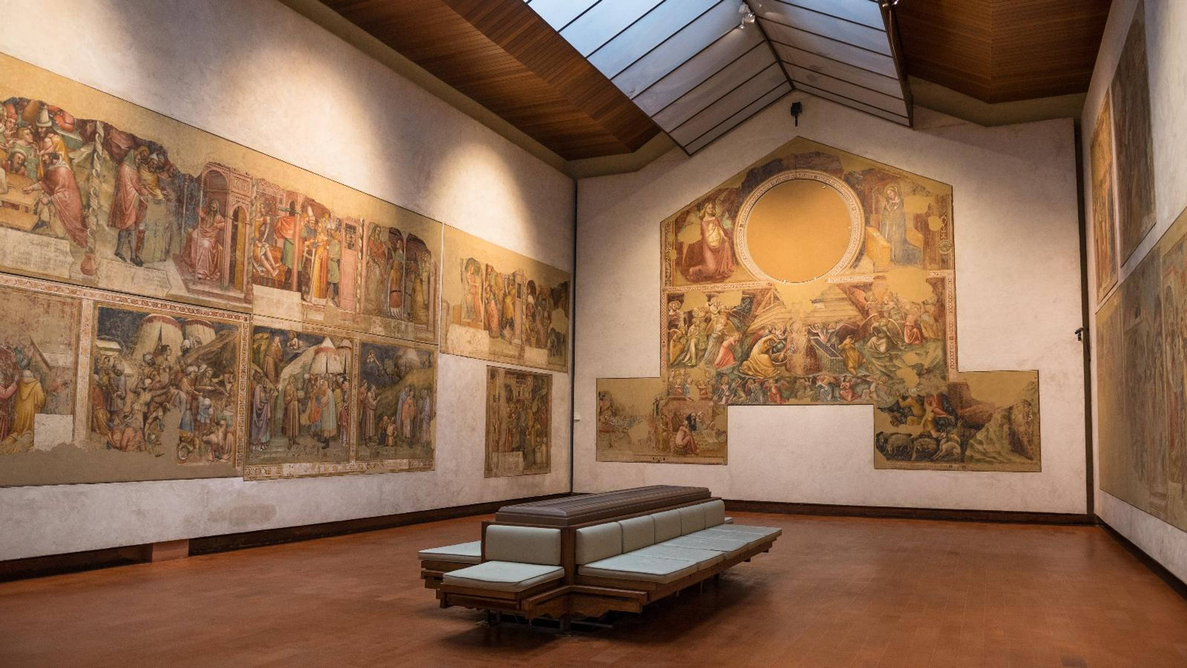 Wnętrze galerii sztuki z rozległymi zabytkowymi freskami na ścianach i ławami ustawionymi pośrodku sali.