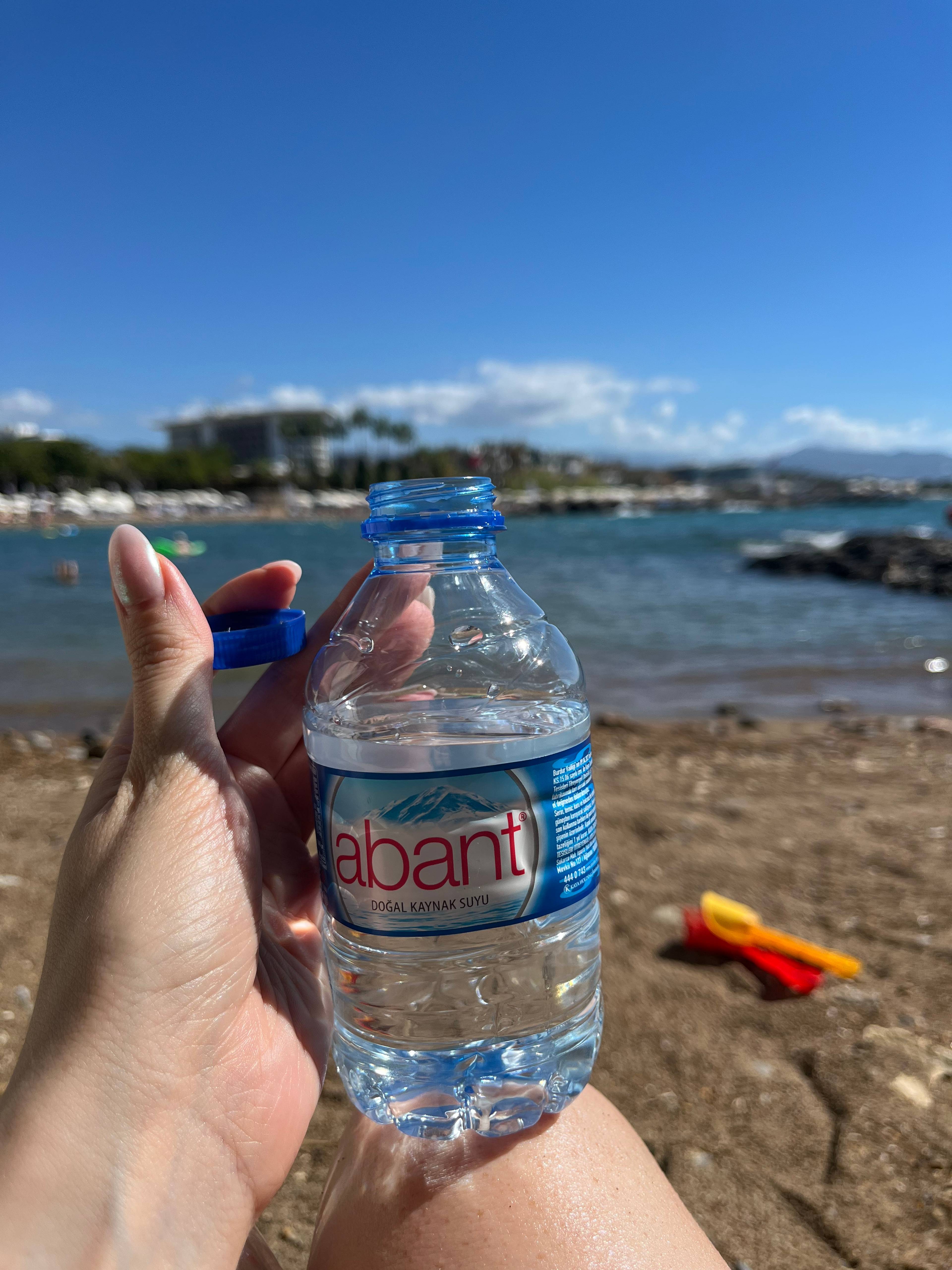 Dłoń trzyma butelkę wody na tle plaży i morza w słoneczny dzień.