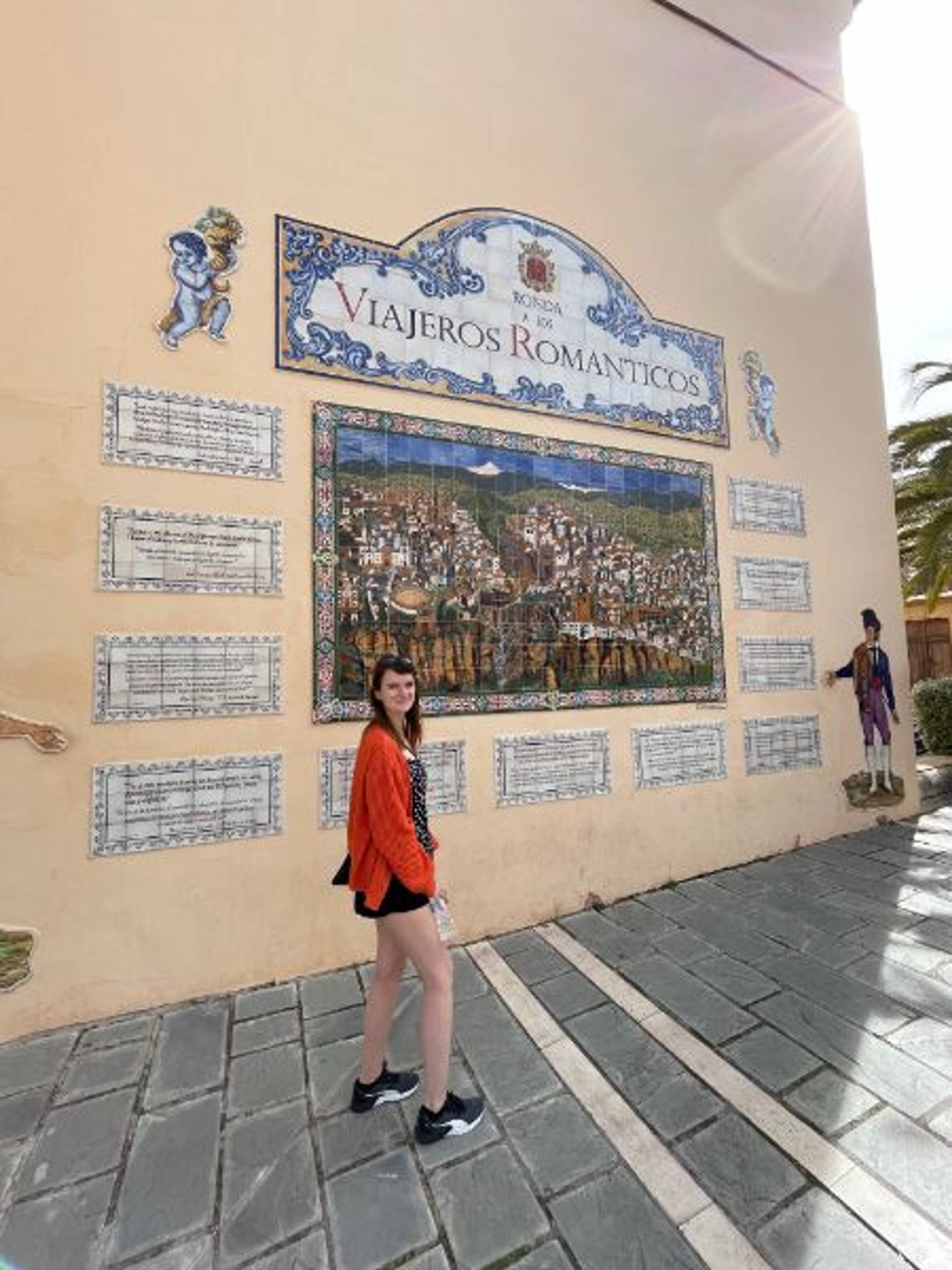 Dziewczyna pozująca przed mozaiką przedstawiającą miasto ronda i napis "Viajeros Romanticos", czyli romantyczne podróże. 