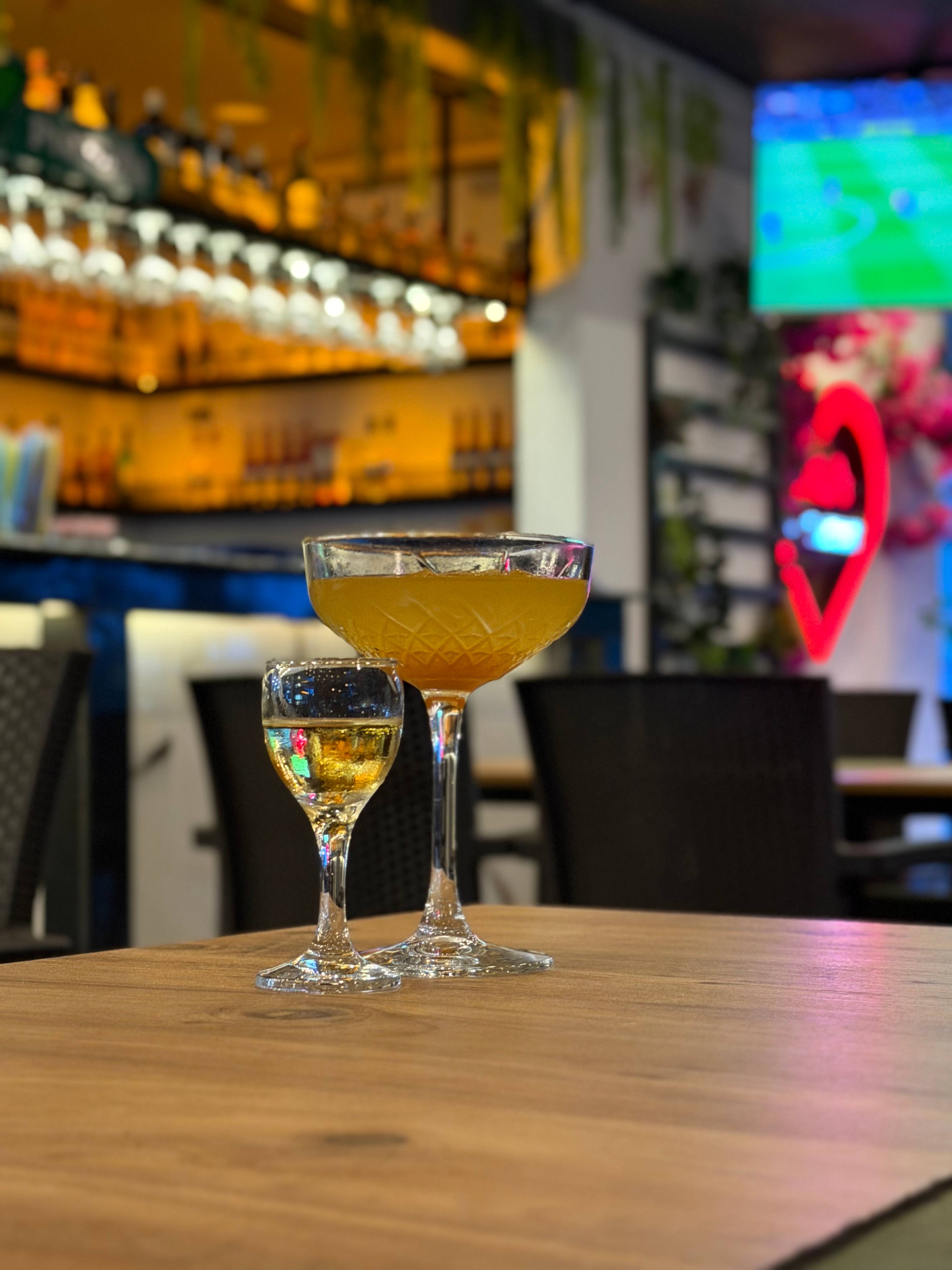 Dwa drinki na drewnianym stoliku w barze, w tle półki z alkoholem i kolorowy ekran.