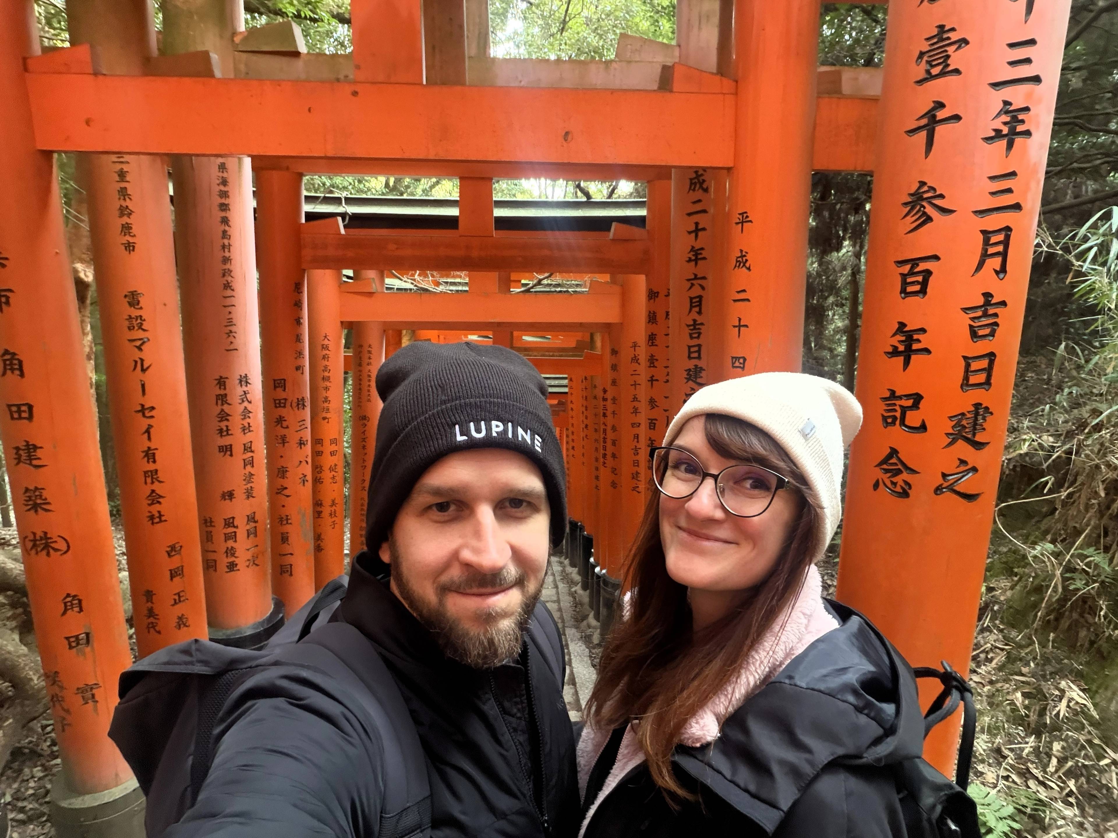 Dwie osoby stoją na ścieżce otoczonej charakterystycznymi pomarańczowymi bramami torii w kompleksie Fushimi Inari w Kioto.