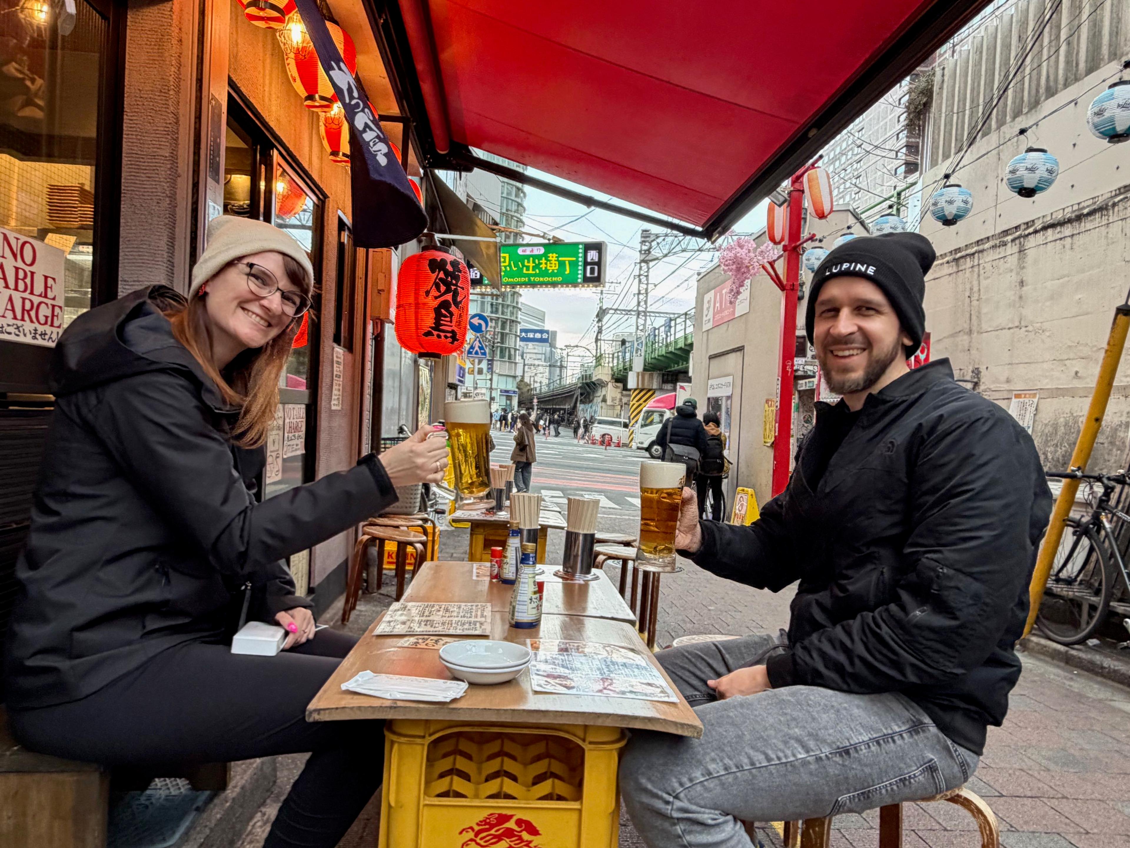 Dwie osoby siedzą przy małym stoliku w ulicznym barze w tokijskiej dzielnicy Omoide Yokocho i wznoszą kufle piwa, a w tle widać japońskie latarnie, szyldy oraz wąską miejską ulicę.