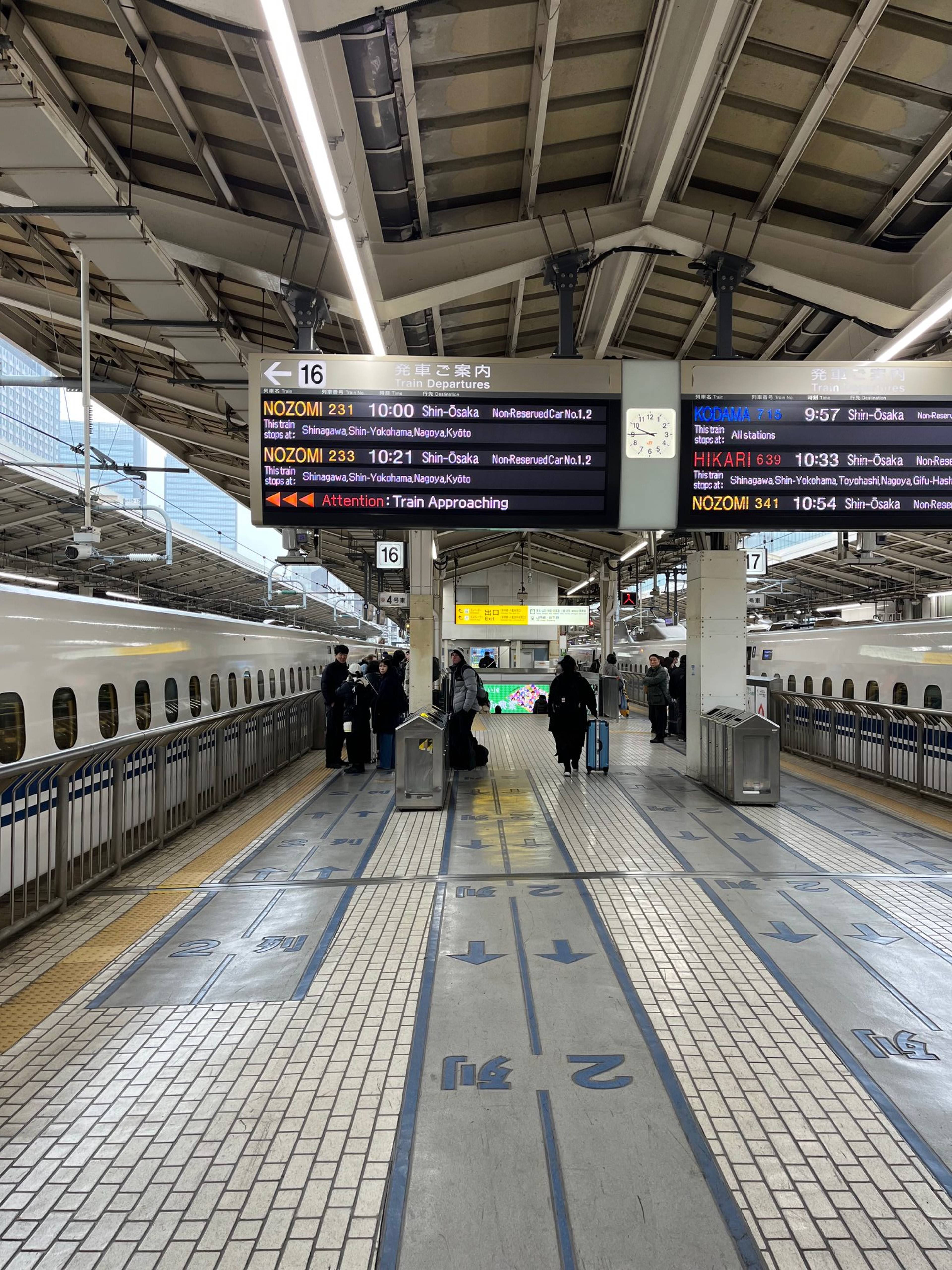Zadaszony peron kolejowy z pociągiem shinkansen po lewej stronie, oznaczeniami torów na posadzce i tablicami odjazdów nad pasażerami.