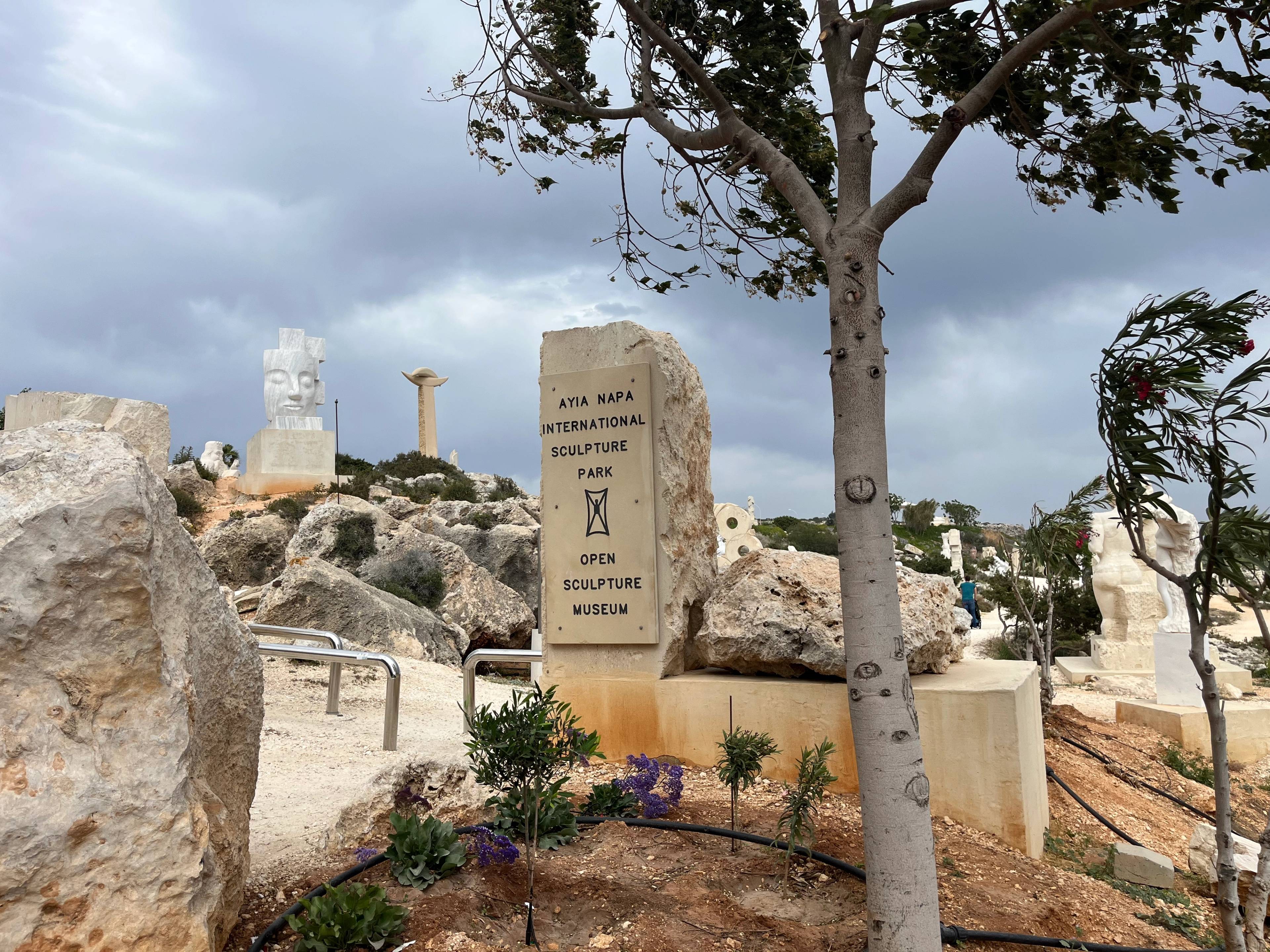 Kamienna tablica z napisem "Ayia Napa International Sculpture Park" w otoczeniu rzeźb na tle skalistego krajobrazu.