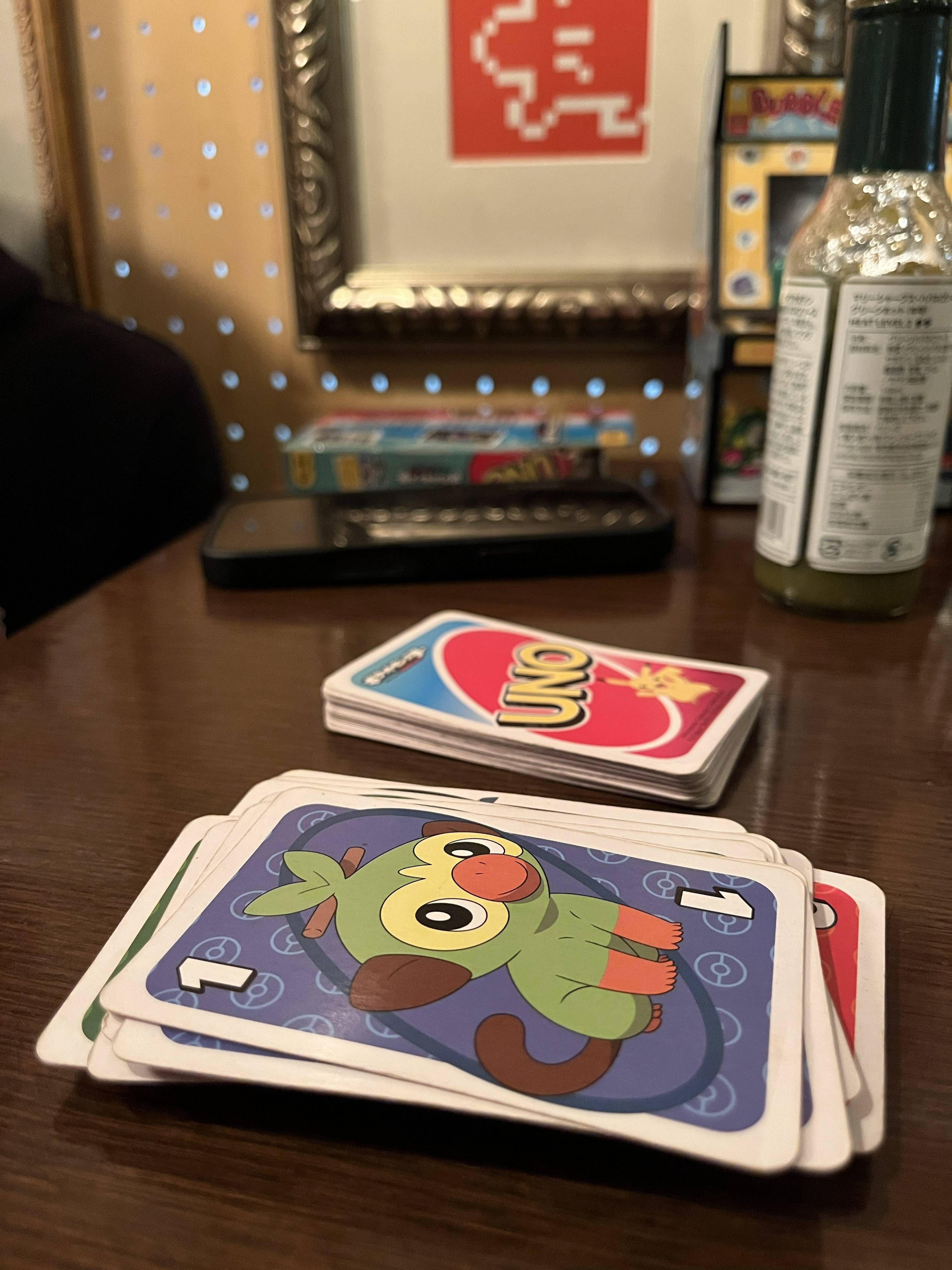 Karty do gry UNO rozłożone na stole podczas rozgrywki, z widoczną kartą przedstawiającą postać Pokémon oraz talią kart w tle.
