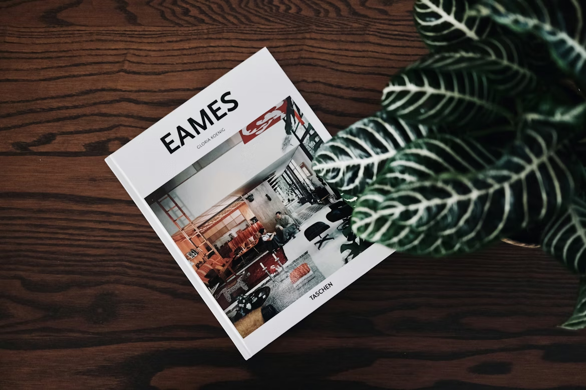 An Eames book on a wooden desk beside a plant