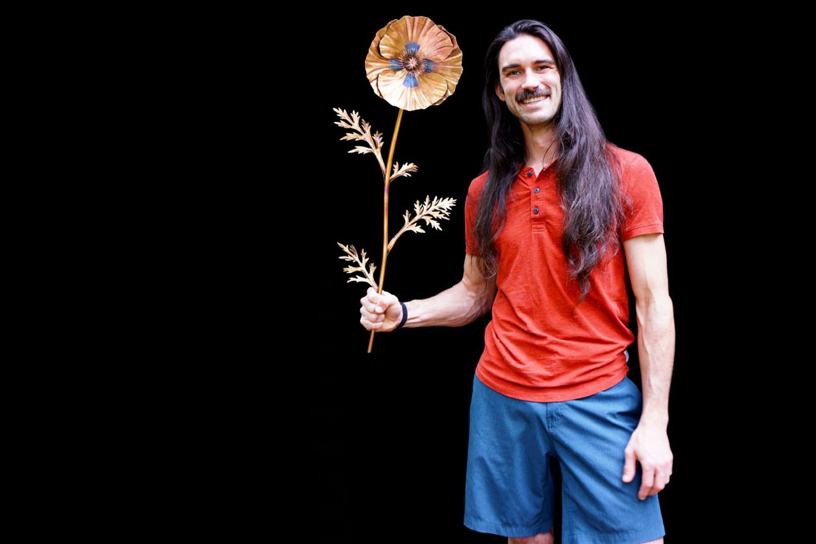 Artist Aiden Dale holding his stainless steel golden poppy sculpture, a handmade flower wall art piece painted only with torch fire.