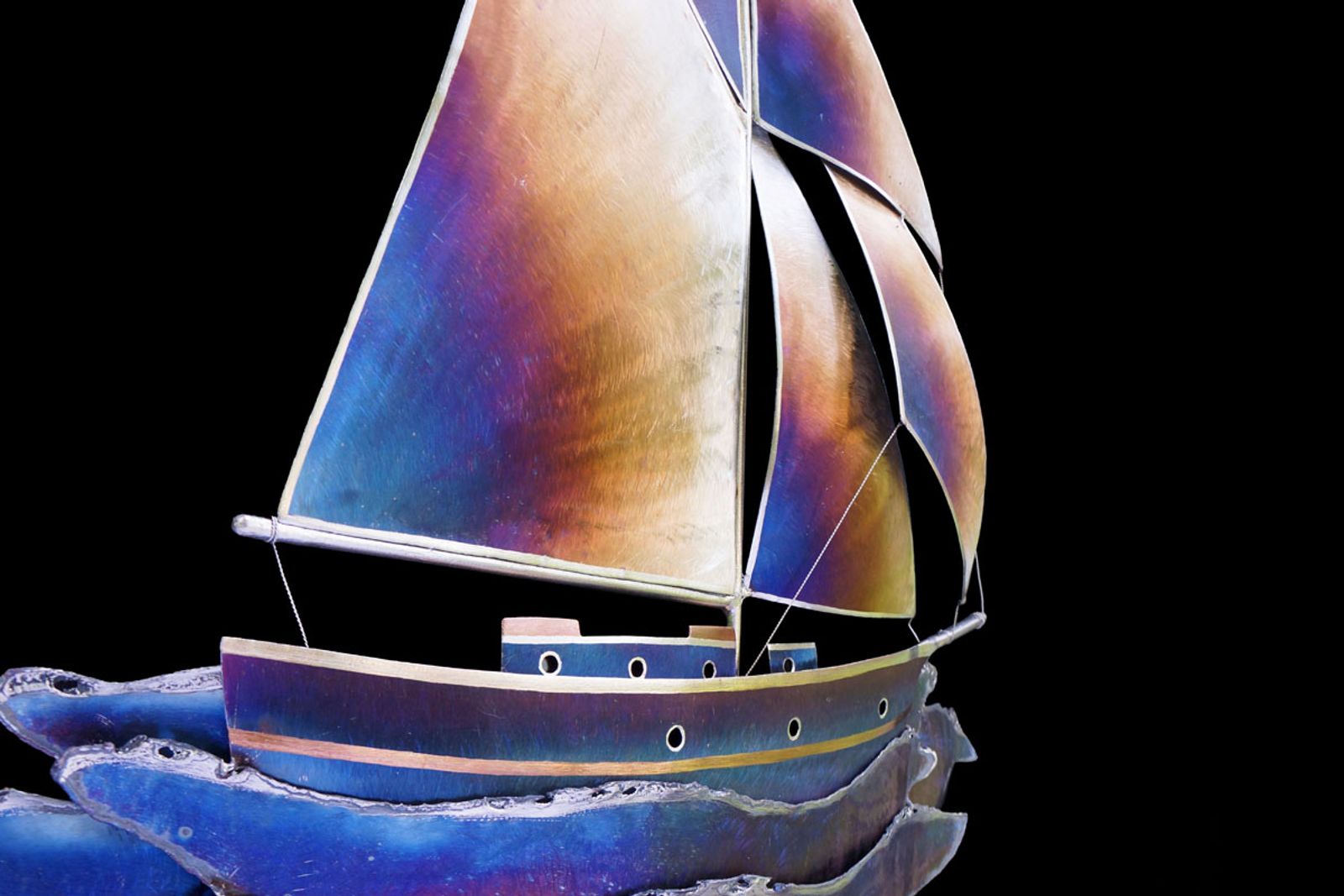 Angled view of stainless steel sailboat wall art showing layered sails, hand colored hull, and blue waves with torch-burned froth.