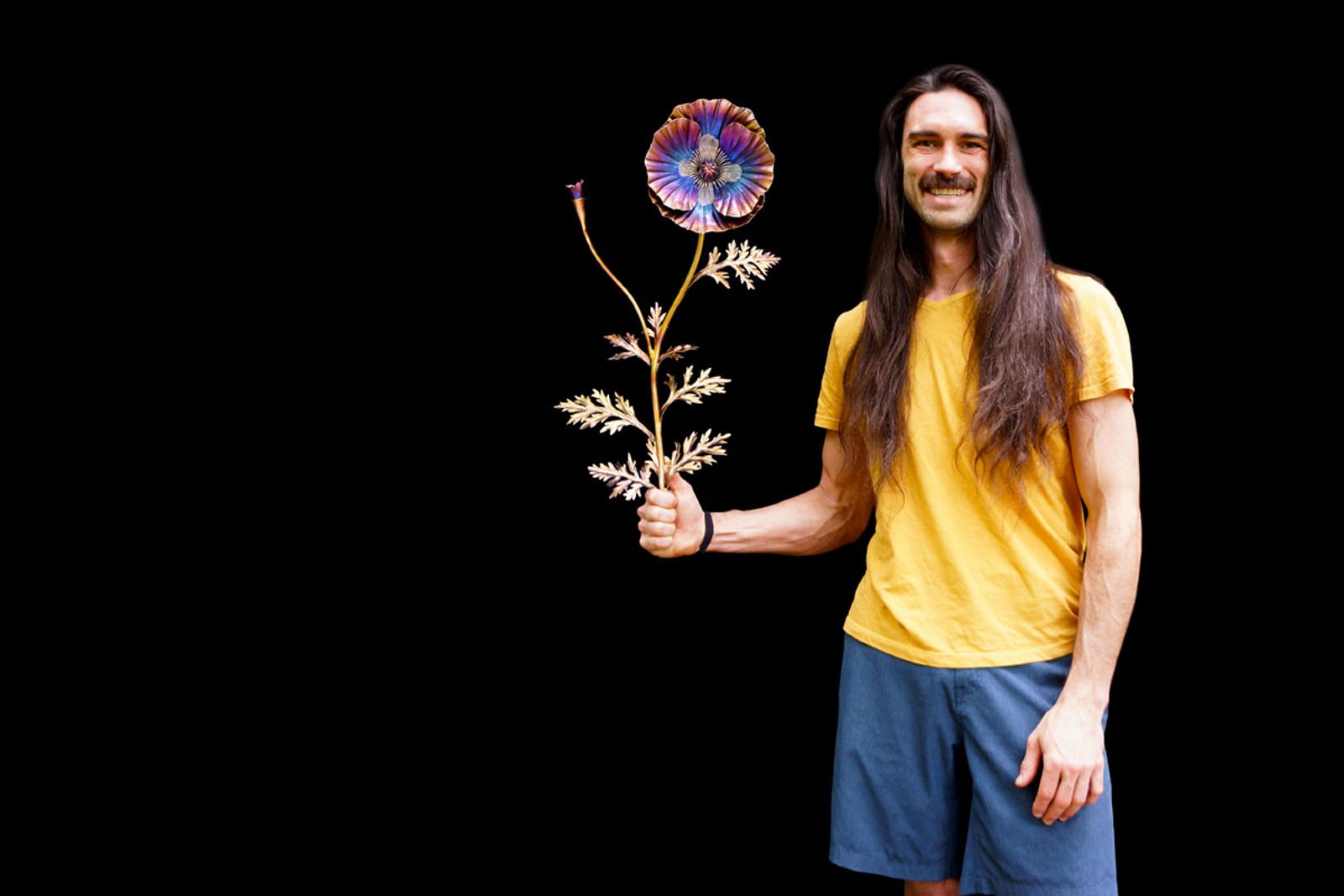 Artist Aiden Dale holding his stainless steel poppy sculpture, handmade with torch patina, intricate stamen bundle, and root system.