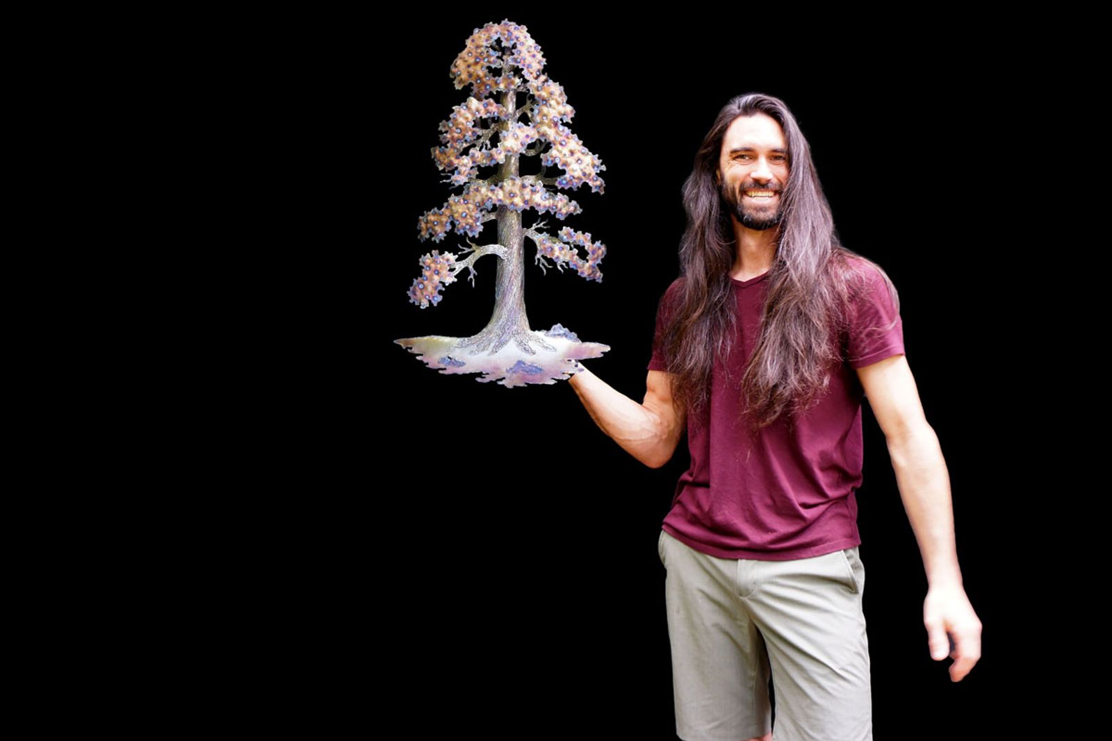 Artist Aiden Dale holding his stainless steel sequoia wall sculpture with layered welded bark and flame-painted foliage