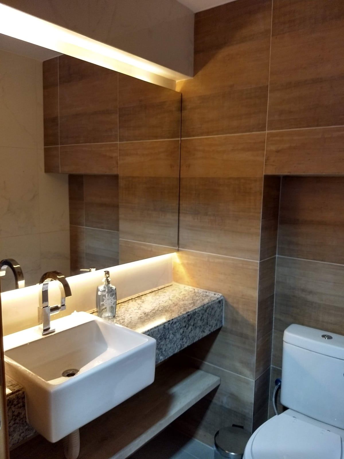 Lavabo com revestimento amadeirado, bancada em granito e espelho com iluminação embutida