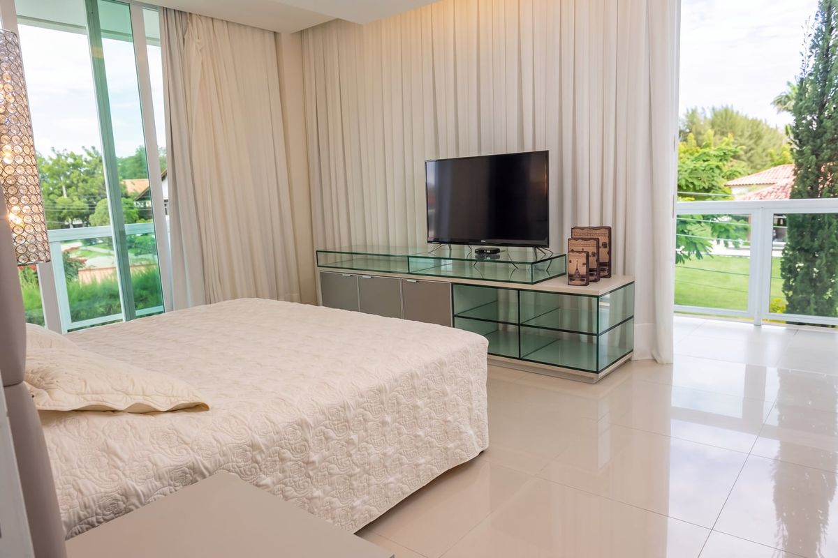 Suíte com marcenaria em vidro, TV e varanda envidraçada voltada ao jardim