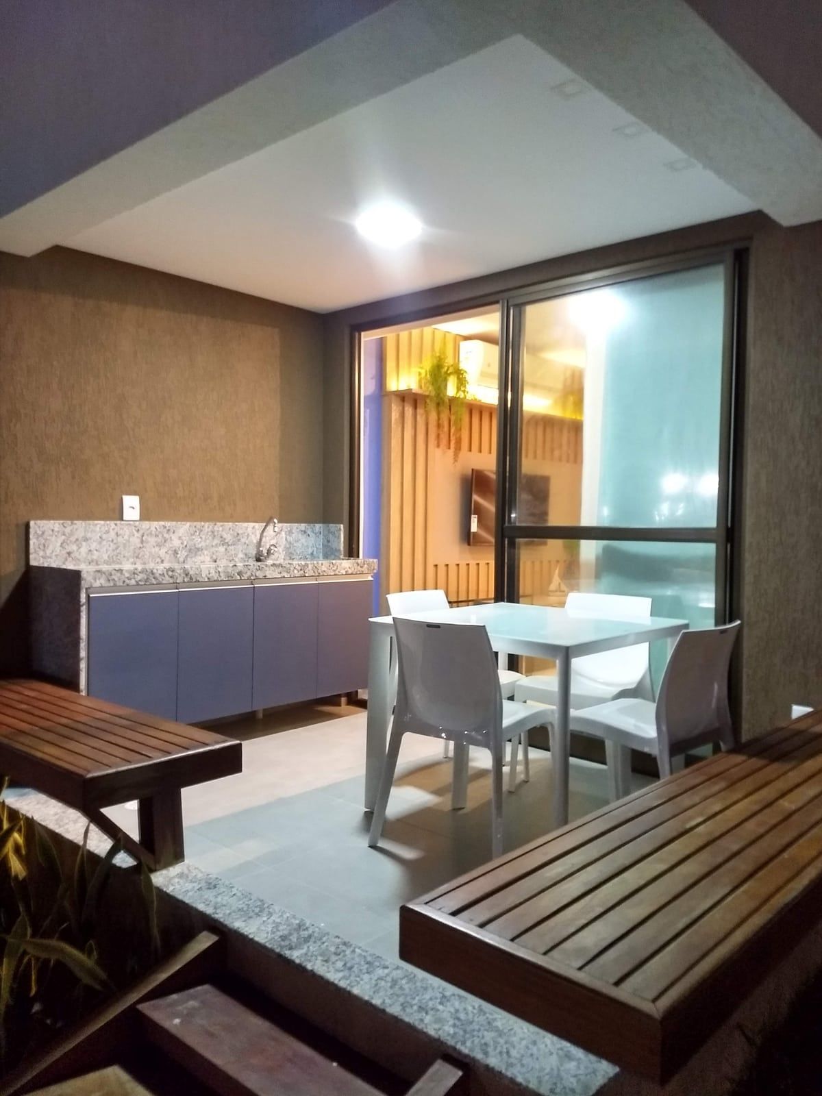 Varanda com bancada gourmet, mesa e bancos, integrada ao interior por porta de vidro