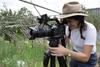 A young woman with her head tilted forward looking into the viewer of a movie camera on a tripid.