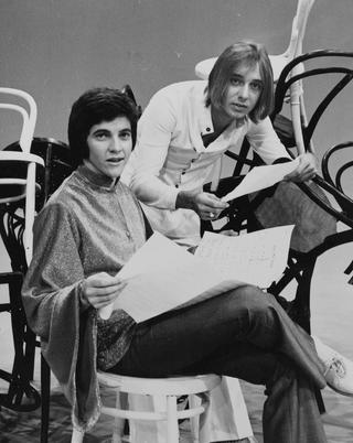 Singer-host Jeff Phillips and singer Russell Morris confer on the set of 1970 pop TV show Sounds Like Us. Phillips is seated and looking over pages of sheet music with Morris leaning on his shoulder. Both are looking at the camera and they are surrounded on the studio floor by a sculptural arrangement of chairs