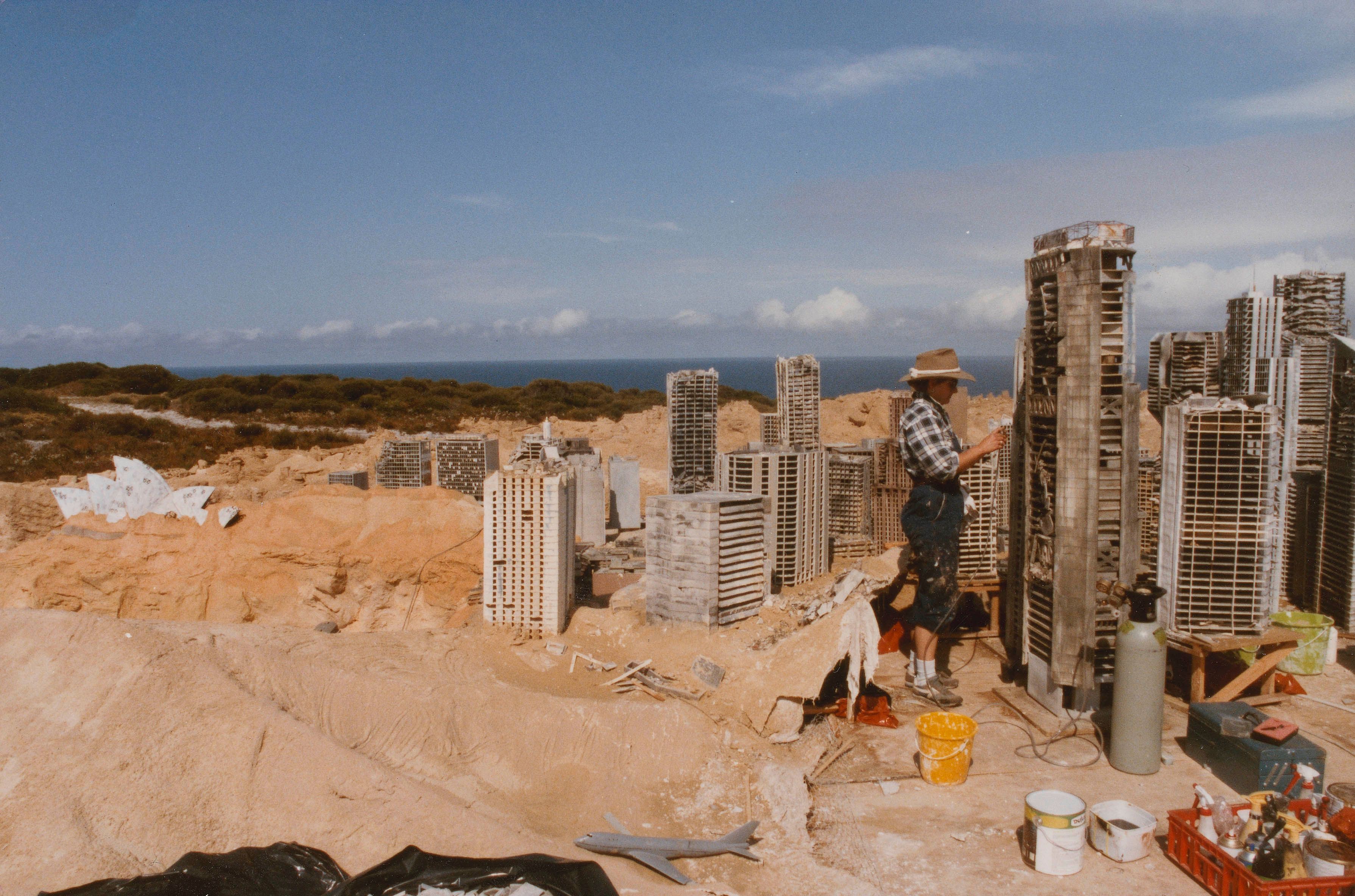 A crew member constructing a model set of post-apocalyptic Sydney in some sand dunes. The buildings are about the size of the person. The Opera House is visible. 