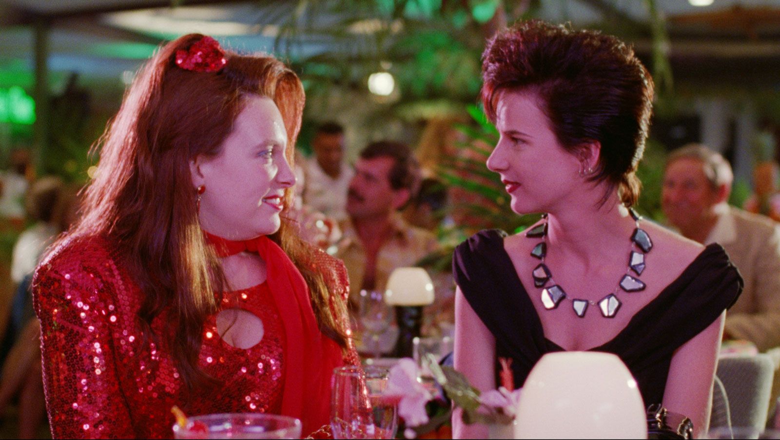 A scene from Muriel's Wedding with Toni Collette and Rachel Griffiths seated and looking directly at each other. They are both dressed up for a night out.