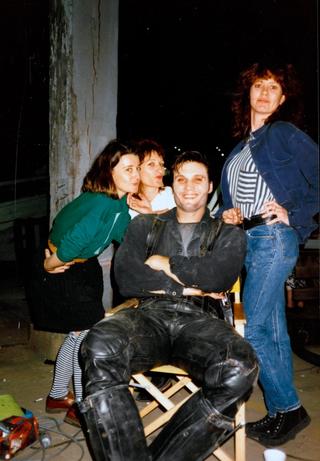 A man sits in a chair dressed in black leather clothing with his arms folded and three woman stand around him.