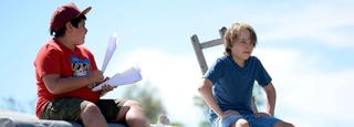 Two boys sit next to each other, with one holding a bunch of paper planes.