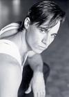 Paul Mercurio in a Strictly Ballroom publicity shot, crouching down on the floor with one elbow resting on his knee and looking sideways towards the camera.