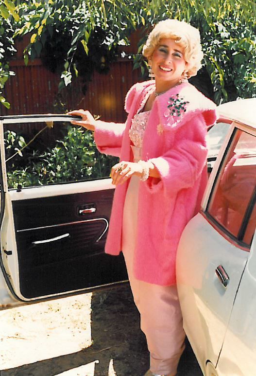 A woman wearing a bright pink jacket standing next to the open passenger door of a car and smiling.