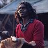 David Dalaithngu as Fingerbone Bill sitting down on the outdoor set of Storm Boy holding a fish.