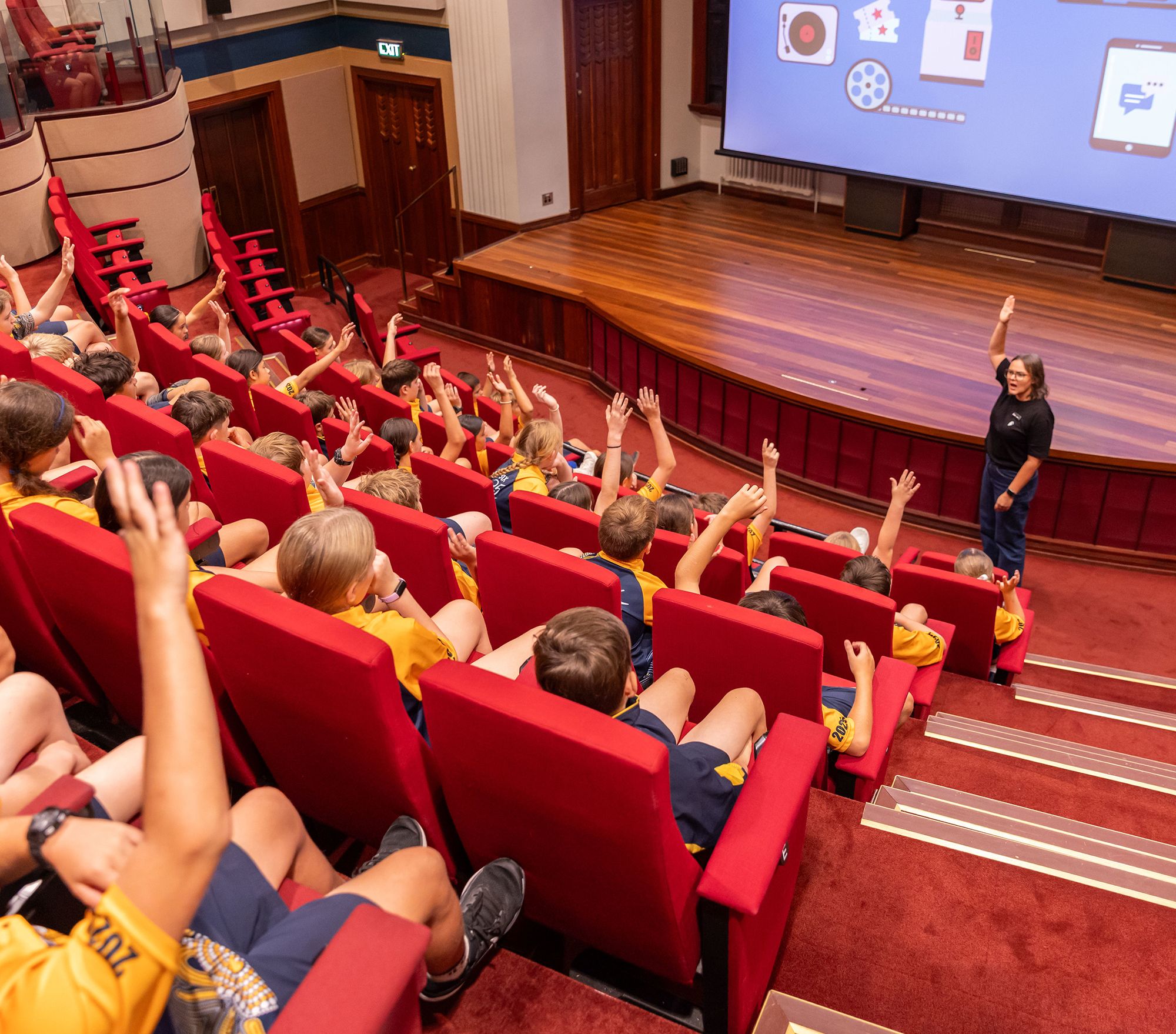 A woman stands at the front of a theatrette packed with school students, many who have a hand raised in the air.