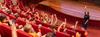 A woman stands in front of a full theatrette of school children, many who have raised one hand in the air to nominate themselves in some way.