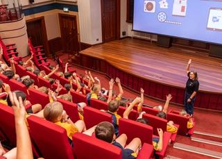 A woman stands at the front of a theatrette packed with school students, many who have a hand raised in the air.