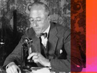 An older man sitting at a desk holding a telephone, c1935