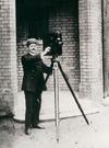 Sidney Cook standing outside a building with his movie camera mounted on a large tripod.