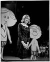 Singer Olivia Newton-John on stage in a TV studio in an episode of the Go Show. She is pictured full length, standing between two lifesize cartoon props of boys wearing short pants.