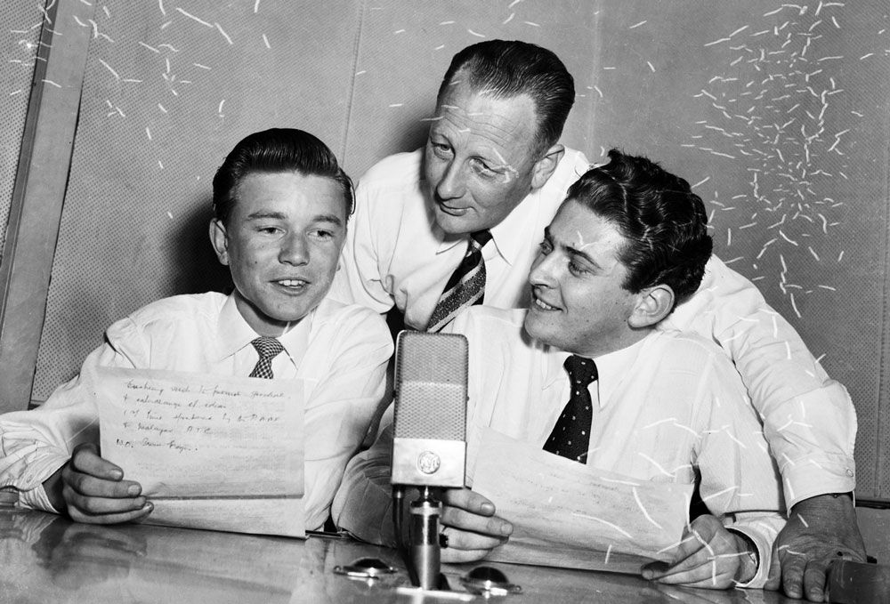 Three radio presenters leaning in towards a microphone, circa 1950s.