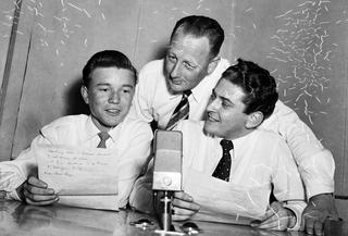 Three radio presenters leaning in towards a microphone, circa 1950s.