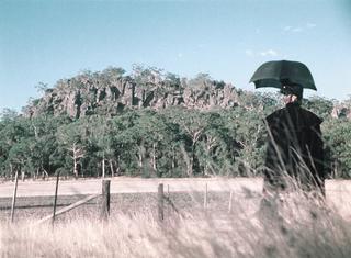 A figure wearing heavy black clothing and holding a parasol stands in a field looking at Hanging Rock rising out of the landscape before them