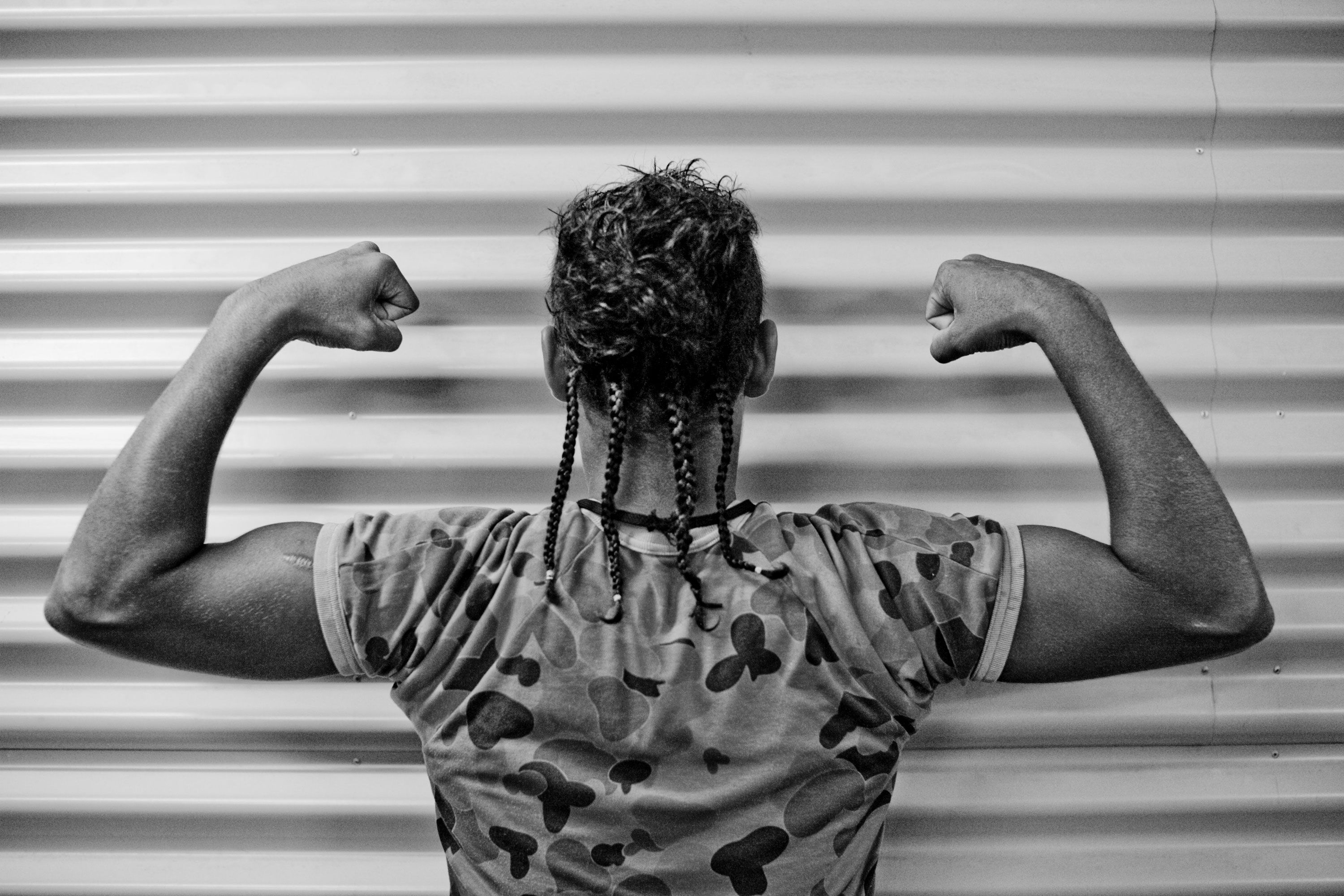 A man standing facing a corrugated iron wall with his back to the camera and arms raised and biceps flexed
