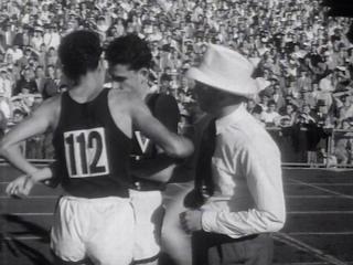 Two athletes and an official on an athletic track with a large crowd behind them in the distance