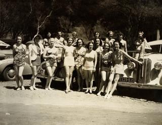Men and woman in swimsuits lined up against cars for a photo.
