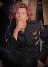 Singer Michael Hutchence kneeling on the ground and resting his chin in his hand and looking directly at camera, wearing a black military style jacket.