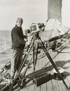 Photo of Frank Hurley on the ship's deck with his camera mounted on a tripod